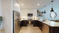 Located at 88 Arsenal St in Watertown, MA, this single-floor residence offers a thoughtfully designed open-concept layout that seamlessly connects the kitchen, dining, and living areas. The kitchen is a standout feature, boasting a U-shaped configuration with two-tone cabinetry — dark lower cabinets paired with crisp white uppers — complemented by a classic white subway tile backsplash, quartz countertops, stainless steel appliances, and open wooden shelving. A kitchen island with a dark base provides additional seating, creating a natural transition into the dining and living spaces. The living area is bright and welcoming, with large windows that invite natural light, light-toned hardwood flooring throughout, and built-in shelving that adds both function and character. The bedroom is a serene retreat, featuring generous natural light from large windows, a spacious open closet, and clean white walls that create a calm ambiance. The bathroom is finished with a walk-in shower clad in white tile, dark cabinetry with gold hardware, and open shelving for storage. Throughout the residence, a cohesive modern aesthetic is maintained with neutral tones, warm wood accents, and contemporary fixtures.