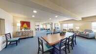 Located at 22000 E Quincy Ave in Aurora, Colorado, this single-floor community space is thoughtfully designed to accommodate a variety of social and functional activities. The layout flows seamlessly across several distinct zones, all unified by soft blue carpeting and warm neutral wall tones throughout.

The main gathering area features a large wooden dining table with padded chairs, complemented by bold artwork including a striking red pear painting. Sliding partitions connect this space to an adjacent multi-purpose room furnished with light-colored tables and modern blue chairs, ideal for meetings or group activities.

A fully equipped kitchen anchors one end of the space, outfitted with dark espresso cabinetry, stainless steel appliances including a range, microwave, and dishwasher, and ample counter space for food preparation and entertaining.

The lounge area offers comfortable seating arrangements, including gray and orange modular chairs paired with a large wall-mounted flat-screen TV, creating a relaxed media viewing environment. Additional seating nooks feature plush armchairs positioned beside wide windows that offer expansive views of the Aurora skyline and surrounding neighborhoods, allowing natural light to fill the space. The overall ambiance is welcoming, functional, and well-suited for community gatherings.