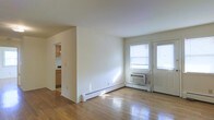 This single-floor space offers a clean, straightforward layout designed for comfortable everyday living. The main living area is finished with warm hardwood floors that reflect natural light streaming in through two windows positioned near the entry door, creating a bright and welcoming atmosphere. The walls are painted in a neutral white tone throughout, contributing to an open and airy feel.

Climate control is provided by a wall-mounted air conditioning unit situated beneath the windows, while baseboard heating runs along the perimeter walls, ensuring year-round comfort. A doorway from the main living area leads directly into the kitchen, which features wood-toned cabinetry and visible countertop space, providing a functional cooking environment. The straightforward flow between the living and kitchen areas supports an efficient use of the available space.

The overall design is simple and practical, with minimal ornamentation, making it a versatile space that can be easily personalized. The combination of hardwood flooring, neutral walls, and natural light gives the interior a clean, livable character suited to a variety of occupants.