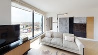 This single-floor residence offers a refined contemporary design with an open, flowing layout. The living room serves as the centerpiece of the space, anchored by oversized floor-to-ceiling windows that flood the interior with natural light and frame expansive city views stretching across the skyline. Light hardwood flooring runs throughout, complementing the neutral palette of whites and warm earth tones. A comfortable sectional sofa, round coffee table, and wall-mounted television furnish the living area, which connects seamlessly to the dining and kitchen zones.

The kitchen features flat-front light wood cabinetry paired with dark stone countertops, stainless steel appliances, and a bold cylindrical pendant light fixture above the parquet-patterned wooden dining table. The design balances warmth and function effectively.

The primary bedroom is generously proportioned, with access to an outdoor-facing window offering city views, a upholstered platform bed, dark wood nightstands, and a built-in closet. A second bedroom features a similar aesthetic with built-in storage and wall sconces. Both rooms maintain a calm, hotel-like ambiance.

The bathroom includes stone-tile flooring, a wall-mounted vanity with a gold-framed mirror, warm sconce lighting, and a full walk-in shower clad in grid mosaic tile.