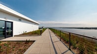 This exceptional rooftop terrace offers a thoughtfully designed outdoor environment that seamlessly blends sustainable landscaping with functional outdoor living. The space is characterized by sweeping green roof sections covered in lush turf and native plantings, interspersed with wide, paved concrete walkways that guide visitors through the area with ease.

A standout feature is the elongated promenade running alongside the building's edge, bordered by a sleek cable and metal railing system that ensures safety while preserving unobstructed sightlines. From this vantage point, expansive panoramic views of a large body of water stretch to the horizon, with a wooded shoreline visible in the distance. On the opposite side, the terrace opens to sweeping city skyline views, with surrounding urban buildings and green tree canopies filling the landscape.

Seating areas with vibrant red outdoor furniture provide inviting spots to rest and take in the views, while low-profile bollard lighting is positioned throughout for use during evening hours. Architectural screening panels offer wind protection in select areas. The green roof sections feature a mix of ornamental grasses, flowering plants, and mulched beds, contributing to the space's ecological character. Overall, the terrace delivers a dynamic, open-air environment that combines sustainability, aesthetic design, and remarkable dual waterfront and cityscape perspectives.