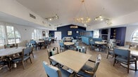 This single-floor community facility offers two distinct, thoughtfully designed areas that cater to both social and wellness needs. The dining and lounge area is an expansive, inviting space anchored by soaring vaulted ceilings and a striking navy blue accent wall that creates a bold focal point. A decorative gold chandelier hangs at the center, complemented by track lighting that illuminates the room with warmth. Multiple dining tables with upholstered blue and patterned chairs are arranged throughout, accommodating a generous number of guests. Arched windows with plantation shutters allow natural light to filter in, while a lounge seating area with plush sofas provides a relaxed gathering space. Artwork and wall décor add character, and hardwood-style flooring ties the aesthetic together seamlessly. The fitness center is outfitted with a comprehensive selection of cardio and strength equipment, including treadmills, rowing machines, stationary bikes, and multi-station weight machines. Blue-painted walls continue the cohesive color theme, and large windows invite natural light into the workout space. Full-length mirrors expand the visual depth of the room, and durable carpet flooring provides comfort underfoot. French doors connect the fitness center to the adjacent spaces, encouraging ease of movement throughout the facility.