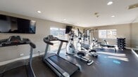 Located at 1147 Briarbrook Dr in Wheaton, Illinois, this fitness center is a well-appointed, single-floor facility designed to support a comprehensive workout experience. The space features durable rubber flooring throughout, recessed lighting, and neutral warm-toned walls that create a clean, focused environment conducive to exercise.

The cardio zone is equipped with multiple treadmills, elliptical trainers, and a stationary bike, all positioned to take advantage of the abundant natural light streaming through large sliding and picture windows that line the walls. Wall-mounted flat-screen televisions provide entertainment during cardio sessions. A Bodycraft rowing machine adds versatility to the cardio offerings.

For strength training, the facility includes a multi-functional cable and weight machine, a weight bench, and a TRX suspension training system anchored to the ceiling, allowing for a wide range of functional fitness exercises. Wooden benches provide seating areas for rest between sets.

A sliding glass door connects the fitness room to an outdoor deck area, offering fresh air and an appealing view of the surrounding landscape. The space also features a mirror-lined wall that visually expands the room and aids in form monitoring during workouts. Motivational signage adorns the walls, contributing to an energizing atmosphere. A door leads to an adjacent room, suggesting additional amenity spaces nearby.