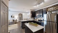 This single-floor residence offers a thoughtfully designed open-concept layout that seamlessly connects the kitchen, dining, and living areas. The kitchen serves as the centerpiece, featuring dark espresso cabinetry with sleek hardware, granite countertops, and a central island that provides generous prep space and casual seating. Stainless steel appliances — including a refrigerator, electric range, microwave, and dishwasher — complement the space, while a white subway tile backsplash adds a clean, contemporary touch. Pendant lighting above the island and a track lighting system illuminate the workspace with both style and function. Crown molding along the upper cabinetry and ceiling adds an elevated architectural detail. The kitchen opens naturally into the dining area, framed by an elegant arched passageway that leads further into the living space. The living area benefits from the open flow, offering warmth and connectivity to the rest of the home. Wood-look plank flooring runs throughout, grounding the space with a cohesive, modern aesthetic. Neutral wall tones provide a versatile backdrop that complements the dark cabinetry and granite surfaces, creating a balanced and inviting atmosphere throughout.