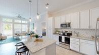 Located at 3839 Galantis Dr in Morehead City, NC, this single-floor residence offers a thoughtfully designed layout with contemporary finishes throughout. The open-concept kitchen serves as the heart of the home, showcasing crisp white shaker cabinetry, a herringbone tile backsplash, granite countertops, and a large center island with seating. Stainless steel appliances, including a refrigerator, range, and microwave, complete the space, while globe pendant lighting adds a stylish touch. Crown molding and light wood-style flooring extend through the main living areas, lending a polished, cohesive aesthetic. The living room is bright and airy, featuring large windows with transom panels above, a ceiling fan, and direct access to an outdoor balcony that frames views of surrounding greenery. Two well-proportioned bedrooms are finished with plush carpet, ceiling fans, and windows that bring in ample natural light. A dedicated bathroom features white subway tile surrounding a tub-shower combination and granite vanity cabinetry. A spacious walk-in closet with wire shelving provides generous storage. A separate laundry area with washer and dryer hookups adds everyday convenience to this well-appointed coastal North Carolina residence.