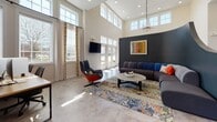 This single-floor community clubhouse in North Chesterfield, Virginia, offers a thoughtfully designed amenity space that balances social gathering, productivity, and fitness. The main lounge area immediately impresses with soaring ceilings, an abundance of natural light streaming through tall windows and transom glass, and a refined yet eclectic décor. A bold dark accent wall, colorful area rug, tufted sectional sofa, and a statement swivel chair in burnt orange create a vibrant yet comfortable atmosphere, complemented by a modern chandelier and wall-mounted television. Large double-glass entry doors open directly to the outdoors, reinforcing the bright and welcoming ambiance throughout. Adjacent to the lounge, a kitchenette features a sleek bar-height counter with teal barstools, pendant lighting, and a natural stone tile accent wall, providing a casual space for refreshments and conversation. A dedicated co-working area offers a communal wood-top table with black seating, ideal for productive gatherings. The expansive fitness center is comprehensively equipped with treadmills, stationary bikes, cable machines, free weights, and strength-training equipment, accented by energetic lime-green walls, floor-to-ceiling mirrors, and ample natural light from multiple windows. A clearly posted fitness center policy and dedicated supply station ensure a well-maintained workout environment throughout.