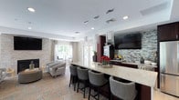 This single-story space offers a thoughtfully designed open-concept layout that seamlessly blends a social bar area with a comfortable lounge. The centerpiece of the space is a generous granite-topped bar counter, accented by modern pendant lighting suspended from the white ceiling above. Upholstered bar stools in neutral gray tones line the counter, providing ample seating for casual gatherings.

Behind the bar, a mosaic tile backsplash adds texture and visual interest, complemented by dark cabinetry and a wall-mounted flat-screen television. A beverage station is conveniently integrated into this area, enhancing the space's functionality.

The adjoining lounge area features plush gray armchairs arranged on a patterned area rug, creating an inviting setting for relaxed conversation. Natural light filters through multiple windows dressed with sheer curtains, contributing to the bright and airy ambiance. French doors lead to an outdoor seating area, further extending the usable space. A stone-textured accent wall adds a distinctive architectural element, rounding out the polished, contemporary aesthetic of this well-appointed communal area.