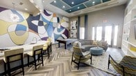 This single-floor lounge space makes a striking first impression with its large-scale abstract mural, which serves as the room's focal point. The mural's sweeping curves and bold palette—featuring deep purples, blues, gold, and white—infuse the space with an artistic, contemporary energy that sets the tone throughout.

The layout is thoughtfully zoned to accommodate multiple uses. A wet bar with a white countertop and bar-height seating provides a social hub for entertaining, while a regulation pool table positioned centrally invites casual recreation. The lounge area features a curved sectional sofa paired with patterned accent chairs in chevron and geometric prints, all arranged around a tufted ottoman and layered area rug for a cohesive, comfortable gathering space.

Overhead, a tray ceiling painted in deep teal adds architectural dimension and contrasts elegantly with the neutral walls. Recessed lighting and track lighting ensure the space is well-illuminated throughout. Herringbone-patterned hardwood flooring runs throughout, adding texture and visual interest underfoot. Natural light filters in through tall windows and glass-paneled doors, creating a bright and welcoming ambiance overall.