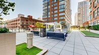 Located at 160 Morgan St in Jersey City, NJ, this rooftop amenity deck offers a well-appointed outdoor retreat set against the backdrop of the surrounding urban skyline. The space is defined by large-format porcelain tile paving that extends across the main terrace, providing a clean, contemporary aesthetic throughout.

Multiple seating arrangements are distributed across the deck, including modern outdoor sofas and lounge chairs in both neutral and navy tones, offering residents ample space to relax and socialize. A white pergola structure serves as a focal architectural element, adding a classic contrast to the modern high-rise surroundings. A swimming pool with surrounding pool deck is also present, complemented by colorful umbrella shading for leisure use.

Stretches of lush artificial turf lawn punctuate the hardscape, including a dedicated bocce court flanked by a high-top bar area with an outdoor television — ideal for casual gatherings. A separate fenced pet area features artificial turf and waste station amenities, designed specifically for residents with dogs.

Landscaped planters with trees and shrubs add greenery throughout the terrace, softening the urban environment. The deck is enclosed by sleek black metal railings that frame open views of the Jersey City skyline. Lighting fixtures and security cameras are integrated throughout, ensuring functionality across all hours.