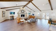This well-appointed single-story space offers a refined yet relaxed atmosphere, highlighted by striking architectural details throughout. The main living area draws immediate attention with its soaring vaulted ceiling adorned with rich, dark-stained exposed wood beams, flooding the space with a sense of volume and warmth. A dramatic floor-to-ceiling stone fireplace serves as the room's centerpiece, flanked by stacked firewood and topped with a circular mirror, creating a bold focal point.

The open layout accommodates multiple seating arrangements, including leather sofas, accent chairs, and a bar-height table with stools, making the space equally suited for casual gatherings or focused work. Wide-plank hardwood flooring flows throughout, contributing to a cohesive, modern-rustic aesthetic.

Natural light pours in through large sliding glass doors and windows, offering views of an outdoor pool area and providing a seamless indoor-outdoor connection. A contemporary chandelier with exposed bulbs adds a touch of elegance overhead.

A private office, accessible through frosted glass French doors, provides a dedicated workspace equipped with a desk and seating. The crisp white walls throughout serve as a neutral canvas, accentuating the bold design elements and artwork displayed across the space. Overall, this space balances comfort, functionality, and sophisticated design.