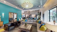 Located at 1502 Partnership Way in Houston, TX, the San Remo clubhouse is a thoughtfully designed single-story community space that blends bold aesthetics with practical amenities. The entrance welcomes visitors through floor-to-ceiling glass doors, offering an immediate view of the resort-style pool beyond, while lush tropical plants and a branded entryway mat set an inviting tone.

The main lounge area is anchored by a large sectional sofa adorned with vibrant teal, blue, and green accent pillows, centered beneath a striking geometric chandelier that casts intricate light patterns across the textured ceiling. Bold teal accent walls serve as backdrops for curated artwork, adding energy to the open, airy interior. The space flows seamlessly into a fully equipped demonstration kitchen featuring dark cabinetry, a sculptural tile backsplash, stainless steel appliances, a generous white quartz island with pendant lighting, and contemporary teal counter stools.

A dedicated media lounge offers a more intimate setting, with a large wall-mounted flat-screen TV, textured tile walls, plush seating, and a gleaming gold ceiling with a sputnik chandelier overhead. The space also includes a private business center or conference area enclosed behind glass-panel sliding doors.

Hexagonal-patterned carpeting in yellow, gray, and neutral tones ties the various zones together, while an abundance of natural light from expansive windows throughout keeps the entire space bright and welcoming.