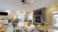 This single-floor space is designed with an open, airy layout that prioritizes comfort and functionality. The main living area is the centerpiece of the space, anchored by a dramatic floor-to-ceiling stacked stone fireplace with a raised hearth and a mounted flat-screen television above the mantel. Vaulted ceilings and recessed lighting enhance the sense of volume, while a ceiling fan adds practical comfort. Light hardwood floors flow throughout, reflecting natural light and contributing to the bright, welcoming atmosphere. Multiple sets of French doors — each topped with transom windows — line the walls, providing abundant natural light and seamless access to an outdoor patio area. The living room is generously sized, accommodating multiple distinct seating arrangements centered around a floral area rug. The adjacent kitchen is well-equipped, featuring cream-colored cabinetry extending to the ceiling, dark granite countertops, stainless steel appliances including a French door refrigerator, and a breakfast bar peninsula ideal for casual dining. Crown molding detail and consistent warm yellow-toned wall colors tie the interior together cohesively. A glimpse into an adjacent room reveals additional entertaining space, suggesting a versatile and well-connected floor plan throughout.