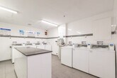This shared laundry facility at 199 Queen Mary Dr in Oakville, Ontario, is a compact yet efficiently organized space designed to serve building residents. The room features a clean, modern aesthetic with white tile walls accented by a horizontal grey tile band, light grey floor tiles, and bright fluorescent ceiling lighting that keeps the space well-illuminated throughout.

The facility is equipped with multiple top-load washing machines arranged along the perimeter walls, as well as stacked washer-dryer units along one wall, providing ample capacity for residents. A payment kiosk is conveniently located within the room, enabling easy card-based transactions for machine use. Instructional signage is posted above the machines to guide users through proper operation.

A central folding counter with a grey laminate surface provides a practical workspace for sorting and folding laundry. A dedicated entry door offers direct access to the room, and a fire safety notice is clearly posted near the entrance. The space also includes a recycling bin and overhead ventilation to maintain air quality. Overall, the facility is thoughtfully laid out to maximize usability within its footprint, offering residents a clean, functional, and organized laundry experience.