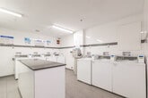 This shared laundry facility at 199 Queen Mary Dr in Oakville, Ontario, is a compact yet efficiently organized space designed to serve building residents. The room features a clean, modern aesthetic with white tile walls accented by a horizontal grey tile band, light grey floor tiles, and bright fluorescent ceiling lighting that keeps the space well-illuminated throughout.

The facility is equipped with multiple top-load washing machines arranged along the perimeter walls, as well as stacked washer-dryer units along one wall, providing ample capacity for residents. A payment kiosk is conveniently located within the room, enabling easy card-based transactions for machine use. Instructional signage is posted above the machines to guide users through proper operation.

A central folding counter with a grey laminate surface provides a practical workspace for sorting and folding laundry. A dedicated entry door offers direct access to the room, and a fire safety notice is clearly posted near the entrance. The space also includes a recycling bin and overhead ventilation to maintain air quality. Overall, the facility is thoughtfully laid out to maximize usability within its footprint, offering residents a clean, functional, and organized laundry experience.