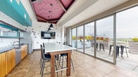 Located in Minneapolis' Uptown neighborhood, this rooftop sky lounge offers a dynamic blend of indoor comfort and outdoor living. The interior is immediately striking, featuring a bold teal accent wall, an intricate herringbone-patterned parquet floor, and a distinctive coffered ceiling clad in rich burgundy wood panels. Sleek black pendant lights hang above a large communal high-top table surrounded by modern bar-height chairs, making it an ideal gathering space. A fully equipped kitchenette lines one wall, showcasing warm wood cabinetry, a scalloped marble tile backsplash, dark granite countertops, a stainless steel sink, ice maker, dishwasher, microwave, and a full-size refrigerator. Decorative bird silhouettes accent the white walls, adding a whimsical artistic touch. Floor-to-ceiling sliding glass doors connect the interior seamlessly to the expansive outdoor terrace. Outside, large-format concrete tiles anchor the space, which is furnished with vibrant teal sectional seating, bistro-style bar tables, and built-in planters overflowing with colorful seasonal flowers. A built-in outdoor grill and glass railing panels complete the rooftop experience, offering sweeping views of the Minneapolis skyline and surrounding neighborhood.