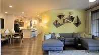 This single-floor residence showcases an open-concept layout that seamlessly connects the living area and kitchen, creating a cohesive and functional flow. The living space is anchored by a plush gray sectional sofa with chaise, positioned beneath a distinctive yellow-green accent wall adorned with geometric, diamond-shaped wooden shelving units that serve as both storage and artistic focal points. A sleek floor lamp provides warm ambient lighting, complementing the recessed ceiling lights overhead. Rich, dark hardwood-style flooring runs throughout, adding warmth and continuity to the space. The kitchen, visible in the background, features a breakfast bar with modern bar stools and pendant lighting, creating an inviting area for casual dining or entertaining. Arched doorways add architectural character, softening the transition between spaces. The overall color palette—warm neutrals punctuated by the vibrant accent wall—creates an energetic yet comfortable atmosphere. Thoughtful décor details, including framed photos and small decorative objects on the shelving, contribute to a personalized, lived-in feel throughout this well-designed single-level home.