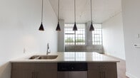Located at 100 Huyshope Ave in Hartford, Connecticut, this loft-style unit showcases a compelling fusion of industrial heritage and contemporary design. The space is defined by its striking oversized factory windows with black steel frames, which flood the interior with abundant natural light and frame views of the surrounding neighborhood. Exposed concrete columns and original masonry along the lower walls pay homage to the building's industrial past, while crisp white walls and soaring ceilings create a bright, airy atmosphere throughout.

Rich hardwood flooring flows seamlessly across the open living areas and bedroom, adding warmth that balances the raw architectural elements. The kitchen is well-appointed with stainless steel appliances — including a refrigerator, range, microwave, and dishwasher — complemented by contemporary cabinetry, a classic white subway tile backsplash, and a light stone countertop island with pendant lighting overhead. The bathroom features neutral tile flooring, a modern vanity, and clean, understated finishes. Tall interior doors with dark wood detailing reinforce the loft aesthetic. The overall ambiance is modern yet character-rich, offering a thoughtfully updated living environment that honors the building's architectural legacy.