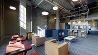 This single-floor space showcases a compelling industrial-modern aesthetic that blends raw architectural elements with refined furnishings. Soaring ceilings with fully exposed ductwork and structural components lend an open, loft-like character to the interior, while pendant lighting fixtures of varying styles hang at different heights to create a warm, layered glow throughout.

A striking stacked-stone accent wall serves as a dramatic focal point, grounding the space and adding rich texture against the smooth, dark-painted surfaces. Large windows along one wall invite abundant natural light, casting dynamic patterns across the striped carpeted flooring.

The lounge is thoughtfully arranged with multiple seating vignettes to accommodate a range of uses. Woven wicker armchairs and deep navy sofas are paired with low coffee tables, encouraging both casual conversation and relaxed lounging. Earthy red upholstered seating adds a pop of color to one corner, enhancing the eclectic warmth of the space.

Toward the rear, a bar-height counter with stools and a refrigerated beverage display hints at a refreshment or social area, making this space well-suited for communal gatherings or events. Structural columns punctuate the floor plan, further reinforcing the industrial character while subtly defining distinct zones within the open layout. Overall, the space balances urban edge with inviting comfort.