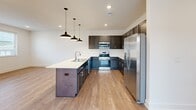 This two-story home offers a well-organized layout across its two levels, with the majority of its living space situated on the upper floor. The entry level features a compact staircase with carpet-covered treads, a sleek metal handrail, and recessed lighting above, creating a clean transition to the main living areas above.

The upper floor houses the open-concept kitchen and living area, where light wood-tone flooring runs throughout. The kitchen is a standout feature, equipped with dark espresso cabinetry, stainless steel appliances including a refrigerator, range, and microwave, and a white countertop peninsula with an undermount sink. Industrial-style pendant lights hang above, adding a modern touch.

The three bedrooms are carpeted and bright, each benefiting from natural light through slatted blinds. The spacious primary bedroom includes a ceiling fan and generous dimensions, while the additional bedrooms offer hardwood-style flooring and exterior views. Each bedroom is served by dedicated closet space.

The two bathrooms feature dark cabinetry vanities with white countertops, large mirrors, and light tile flooring. A dedicated laundry room with a full-size top-loading washer and dryer and overhead wire shelving completes the functional layout.