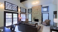This single-floor home makes an immediate impression with its dramatic architectural design and thoughtfully curated details. The living area commands attention with soaring vaulted ceilings accented by dark exposed beams, creating a sense of grandeur and openness. A floor-to-ceiling stacked stone fireplace serves as the undeniable centerpiece of the room, anchoring the space with rustic elegance and warmth.

Large dark-framed windows, including transom windows positioned high along the vaulted walls, flood the interior with generous natural light while framing views of the surrounding outdoor greenery. French doors provide seamless access to an outdoor deck, further enhancing the connection between interior and exterior living. The warm, neutral palette of the walls complements the rich tones of the stone and woodwork throughout.

The living room is furnished with plush leather seating and striped accent chairs arranged around a central coffee table, all resting on a classic area rug that defines the space. Track lighting overhead adds a layer of ambient illumination to complement the natural light. The overall ambiance is one of refined comfort — blending rustic architectural character with a welcoming, livable warmth ideal for both relaxing and entertaining.