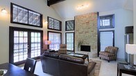 This single-floor home makes an immediate impression with its dramatic architectural design and thoughtfully curated details. The living area commands attention with soaring vaulted ceilings accented by dark exposed beams, creating a sense of grandeur and openness. A floor-to-ceiling stacked stone fireplace serves as the undeniable centerpiece of the room, anchoring the space with rustic elegance and warmth.

Large dark-framed windows, including transom windows positioned high along the vaulted walls, flood the interior with generous natural light while framing views of the surrounding outdoor greenery. French doors provide seamless access to an outdoor deck, further enhancing the connection between interior and exterior living. The warm, neutral palette of the walls complements the rich tones of the stone and woodwork throughout.

The living room is furnished with plush leather seating and striped accent chairs arranged around a central coffee table, all resting on a classic area rug that defines the space. Track lighting overhead adds a layer of ambient illumination to complement the natural light. The overall ambiance is one of refined comfort — blending rustic architectural character with a welcoming, livable warmth ideal for both relaxing and entertaining.