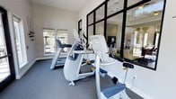 This single-story fitness center is designed to provide a functional and inviting workout environment. The space is outfitted with a selection of modern cardio equipment, including treadmills, elliptical trainers, and stationary bikes, all arranged to maximize usability and floor space. The flooring consists of durable, cushioned grey carpet well-suited for a high-traffic exercise environment.

Natural light fills the room through multiple windows fitted with roller shades, offering views of the surrounding outdoor greenery and contributing to an energizing atmosphere. The crisp white walls and recessed ceiling lighting further enhance the brightness of the space.

A standout architectural feature is the bold black-framed glass partition wall, which visually connects the fitness area to an adjacent room—likely a lounge or common area—creating a sense of openness and continuity throughout the facility. Large mirrors are incorporated into the design, amplifying the sense of space and allowing users to monitor their form during workouts.

Overall, the fitness center strikes a balance between practicality and modern aesthetic appeal, offering a clean, well-lit, and thoughtfully designed space for exercise.