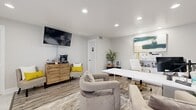 This thoughtfully designed single-floor office space offers a seamless blend of professional functionality and modern aesthetics. The layout is divided into two distinct zones: a reception and lounge area near the entrance, and a dedicated workstation area toward the rear.

The entry zone features large-format marble-look tile flooring, a bold gray board-and-batten accent wall, and a glazed entry door flanked by a window with white blinds that allows natural light to filter in. This area transitions smoothly into the main workspace, where durable wood-look plank flooring and a patterned area rug add warmth and texture.

The workstation area is anchored by a sleek white desk paired with a white and gold executive chair, complemented by a dual-monitor setup and organized shelving. Plush, neutral-toned swivel chairs are arranged to accommodate client consultations, while a small side table adds convenience. A vibrant abstract canvas artwork above the desk adds a sophisticated, creative touch.

A wall-mounted flat-screen television and a wood-paneled coffee station with two accent chairs with yellow cushions provide a comfortable, welcoming atmosphere for visitors. Recessed lighting throughout ensures bright, even illumination across the entire space. Potted greenery and decorative accents complete the polished, professional ambiance.