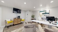 This thoughtfully designed single-floor office space offers a seamless blend of professional functionality and modern aesthetics. The layout is divided into two distinct zones: a reception and lounge area near the entrance, and a dedicated workstation area toward the rear.

The entry zone features large-format marble-look tile flooring, a bold gray board-and-batten accent wall, and a glazed entry door flanked by a window with white blinds that allows natural light to filter in. This area transitions smoothly into the main workspace, where durable wood-look plank flooring and a patterned area rug add warmth and texture.

The workstation area is anchored by a sleek white desk paired with a white and gold executive chair, complemented by a dual-monitor setup and organized shelving. Plush, neutral-toned swivel chairs are arranged to accommodate client consultations, while a small side table adds convenience. A vibrant abstract canvas artwork above the desk adds a sophisticated, creative touch.

A wall-mounted flat-screen television and a wood-paneled coffee station with two accent chairs with yellow cushions provide a comfortable, welcoming atmosphere for visitors. Recessed lighting throughout ensures bright, even illumination across the entire space. Potted greenery and decorative accents complete the polished, professional ambiance.