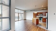 This well-appointed one-bedroom, two-bathroom residence offers a thoughtfully designed open-concept floor plan with a generous overall footprint. The combined kitchen and living area serves as the heart of the home, characterized by rich hardwood-style flooring, soaring ceilings, and an exposed metal ductwork detail that lends an industrial-chic aesthetic. Floor-to-ceiling windows and a glass-paneled balcony door flood the living area with abundant natural light while providing outdoor access and open views.

The kitchen is a standout feature, showcasing warm wood cabinetry, subway tile backsplash, granite-topped counters, and a curved island with pendant lighting overhead. Stainless steel appliances, including a range, microwave, and refrigerator, complete this functional and stylish space.

The bedroom features wood-style flooring, a large window with exterior views, and a well-organized walk-in closet equipped with built-in wire shelving and carpeted flooring for added comfort. Two bathrooms serve the residence, with one featuring a walk-in shower, wood-toned vanity, and granite countertop. A dedicated hallway connects the living spaces efficiently, contributing to a cohesive and practical layout throughout.