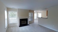 This single-floor residence offers a welcoming and well-lit interior characterized by a clean, open layout and neutral warm tones throughout. The main living area is anchored by a classic wood-mantled fireplace with a black surround and hearth, serving as a natural focal point for the room. Soft, wall-to-wall carpeting extends across the space, adding warmth and comfort underfoot. Multiple double-hung windows flood the interior with abundant natural light, including views of greenery outside, creating a bright and airy atmosphere. The living space flows seamlessly into an adjacent dining or breakfast area, distinguished by a stylish pendant light fixture and a raised countertop bar or pass-through that hints at a connected kitchen beyond. The vaulted or raised ceiling in the adjoining area adds a sense of openness and dimension. The interior is finished with crisp white trim, clean baseboards, and warm beige walls, lending the space a classic, move-in-ready aesthetic. Overall, the home presents a functional and comfortable single-level layout, well-suited for everyday living with a balance of traditional design elements and practical open-concept flow.