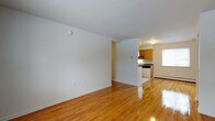 This single-floor, 1-bedroom, 1-bathroom residence offers a clean and functional layout designed for comfortable everyday living. The open living area is bright and welcoming, showcasing polished hardwood floors that run throughout the space, reflecting natural light and adding warmth to the interior. Crisp white walls provide a neutral backdrop, creating an airy and versatile environment that is easy to personalize.

The living area flows directly toward the kitchen, which is partially separated by a structural dividing wall that adds a sense of definition between the two spaces. The kitchen features warm wood cabinetry providing ample storage, a dark countertop workspace, and standard appliances including a range. A window in the kitchen area allows natural light to brighten the room.

A baseboard heating system is visible along the walls, ensuring year-round comfort. The overall design of the space is straightforward and practical, with well-maintained finishes throughout. The combination of hardwood flooring, neutral tones, and a connected kitchen-living layout makes this a highly functional single-floor residence suited for a range of lifestyles.