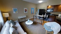 This single-floor space offers a thoughtfully designed open-concept layout that blends the living and kitchen areas into one cohesive environment. The living area is anchored by a comfortable gray sectional sofa paired with a sleek round white coffee table, resting atop a patterned area rug that adds texture and warmth to the space. A geometric console table lines one wall, complementing the contemporary aesthetic throughout. Three framed abstract artworks adorn the warm taupe-toned walls, adding pops of color and personality to the room. The transition into the kitchen is natural and uninterrupted, where a high-top dining table with bar stools serves as both a casual dining and gathering spot. The kitchen is equipped with black appliances, including a full-size refrigerator and microwave, alongside upper cabinetry finished in warm wood tones. Hardwood flooring in the kitchen area provides a clean contrast to the carpeted living space. The overall ambiance is modern yet inviting, with neutral wall tones, curated decor, and a functional layout that makes efficient use of the available space, ideal for comfortable everyday living.