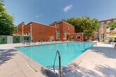 This outdoor amenity space centers around a large, rectangular swimming pool with crystal-clear turquoise water, framed by a spacious tiled sun deck. The pool accommodates varying depths, ranging from a shallow entry zone to deeper sections, with clearly marked depth indicators and convenient metal entry railings. Surrounding the pool, the generous deck is outfitted with an array of blue lounge chairs and colorful umbrellas in shades of green, orange, and yellow, providing ample space for sunbathing and relaxation. One side of the pool area is anchored by a commanding historic red brick building, featuring large arched and multi-pane windows that reflect the structure's industrial architectural heritage. The building's warm, weathered brick facade adds significant character to the outdoor environment. On the opposite perimeter, a contemporary fence lined with vertical paneling and lush greenery panels creates a defined, private boundary. Mature deciduous trees tower above the space, offering natural shade and a lush green backdrop against the open sky. Additional seating nooks with lounge sofas and accent tables are thoughtfully arranged along the fence line, creating semi-private social areas. The overall ambiance blends historic industrial character with vibrant, resort-style outdoor living.