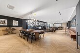 Located at 2946 Dundas St W in Toronto's vibrant Junction neighbourhood, this single-floor lounge space offers a refined and thoughtfully designed interior that blends contemporary aesthetics with warm, inviting elements. The expansive open-plan layout is unified by striking light oak herringbone flooring, which adds texture and visual continuity throughout the space.

The main lounge area is anchored by plush tufted sofas and stylish armchairs arranged around elegant glass and metal coffee tables, creating comfortable conversation zones. A dramatic floor-to-ceiling teal shelving wall, adorned with curated decorative objects, serves as a bold focal point and draws the eye toward large windows that flood the space with natural light.

A dedicated bar area with counter seating provides a functional social hub, while a long communal dining table with pendant globe lighting overhead offers a versatile space for group gatherings. Built-in bench seating along the deep navy painted brick walls, paired with round bistro tables, adds additional informal dining options.

Framed artwork displayed throughout contributes to the space's cultivated, gallery-like character. Recessed ceiling lighting complements the natural light, ensuring the space remains bright and welcoming throughout the day. The overall ambiance is sophisticated yet relaxed, making it an ideal venue for social interaction and community engagement.
