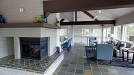 This inviting single-floor space makes a strong architectural impression with its soaring vaulted ceilings adorned by bold, dark wood exposed beams that draw the eye upward and add dramatic contrast to the crisp white walls. At the heart of the room stands a painted white brick fireplace, a true focal point, featuring a raised hearth, a generous mantle shelf styled with decorative glass vessels, and a modern multi-sided glass firebox that adds warmth and visual interest from multiple angles. The flooring throughout is composed of natural slate tiles in an earthy, varied palette, lending texture and a grounded, organic feel to the interior. The open layout flows seamlessly between a casual lounge area, furnished with tufted blue armchairs and a plush sectional, and a bar-height counter with coordinating blue upholstered stools — ideal for both relaxed gatherings and casual dining. Pendant lighting hangs gracefully from the beamed ceiling, complementing the natural light that pours in through large windows along the curved perimeter wall. A sleek metal railing defines the space's open edge, hinting at elevated outdoor views of surrounding greenery, further enhancing the bright and airy ambiance of this thoughtfully designed interior.