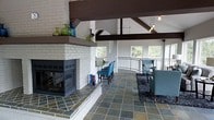 This inviting single-floor space makes a strong architectural impression with its soaring vaulted ceilings adorned by bold, dark wood exposed beams that draw the eye upward and add dramatic contrast to the crisp white walls. At the heart of the room stands a painted white brick fireplace, a true focal point, featuring a raised hearth, a generous mantle shelf styled with decorative glass vessels, and a modern multi-sided glass firebox that adds warmth and visual interest from multiple angles. The flooring throughout is composed of natural slate tiles in an earthy, varied palette, lending texture and a grounded, organic feel to the interior. The open layout flows seamlessly between a casual lounge area, furnished with tufted blue armchairs and a plush sectional, and a bar-height counter with coordinating blue upholstered stools — ideal for both relaxed gatherings and casual dining. Pendant lighting hangs gracefully from the beamed ceiling, complementing the natural light that pours in through large windows along the curved perimeter wall. A sleek metal railing defines the space's open edge, hinting at elevated outdoor views of surrounding greenery, further enhancing the bright and airy ambiance of this thoughtfully designed interior.