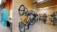 This single-floor facility is purpose-built for cyclists, offering a well-organized and functional space divided into two key areas: a bike storage room and an adjacent locker corridor. The bike storage area is expansive, with plywood-paneled walls lining the perimeter and serving as the mounting surface for a series of wall-mounted vertical bike racks. These racks accommodate a wide variety of bicycle types — from mountain bikes and road bikes to folding bikes — keeping the polished concrete floor clear and maximizing storage capacity. Exposed ceiling infrastructure, including conduit and fluorescent tube lighting, gives the space an industrial character. Large windows along one wall allow natural light to filter in, offering views of a landscaped outdoor area. The adjoining locker corridor features a row of teal-colored metal lockers set against a bold orange accent wall, providing secure personal storage for cyclists. A built-in countertop with cabinetry beneath offers additional utility space. Windows along the corridor continue the theme of natural light throughout. The overall design is practical and efficient, combining durable materials with thoughtful amenities tailored to an active, cycling-oriented user base.