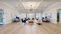 This well-appointed leasing office and business center spans a single floor, offering a thoughtfully designed layout across three distinct areas. The centerpiece is a grand open reception lounge showcasing soaring coffered ceilings, a striking sculptural sputnik chandelier, and light-toned hardwood flooring that flows throughout. Floor-to-ceiling French doors and transom windows flood the space with abundant natural light while offering outdoor views, creating an airy, welcoming atmosphere. The lounge features a curated seating arrangement with tufted navy velvet armchairs, a cognac leather sofa, and a richly textured drum coffee table, anchored by a vibrant abstract area rug. Flanking the lounge are dedicated leasing workstations with warm wood desks, ergonomic seating, and large-scale abstract artwork, providing a professional yet inviting environment for consultations. A private conference and business center room offers a more intimate setting, distinguished by a calming teal accent wall, plush carpeted flooring, a round glass-top table with upholstered blue chairs, and a separate writing desk — ideal for private meetings or focused work. The entry foyer, framed by elegant double doors with decorative ironwork, provides a polished first impression and connects seamlessly to adjacent amenity spaces.