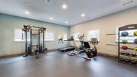 Located at 8855 McGallion Rd in Houston, TX, this fitness center offers a single-floor exercise room designed to support a well-rounded workout routine. The space is thoughtfully laid out to maximize functionality, featuring durable dark rubber flooring that provides both cushioning and stability during exercise.

The room is equipped with a comprehensive selection of fitness equipment, including a treadmill, elliptical trainer, stationary recumbent bike, and a multi-function cable and weight machine for strength training. A dedicated dumbbell rack with a full set of free weights adds further versatility for resistance training. Wall-mounted mirrors allow users to monitor their form throughout workouts.

Natural light filters in through multiple windows fitted with roller shades, creating a bright and energizing environment. Recessed ceiling lighting provides consistent illumination throughout the space. The walls are finished in a clean two-tone palette of soft sage green and warm white, contributing to a calm, motivating atmosphere.

Access to the fitness center is provided through glass-panel doors, and safety features include clearly marked emergency exits and a fire extinguisher. The open floor plan allows for comfortable movement between equipment, making this facility a practical and well-appointed option for residents in the Houston area.