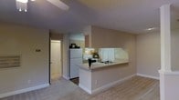 This single-floor space showcases a practical and open layout that connects the kitchen and living area seamlessly. The kitchen is equipped with white appliances, including a refrigerator and range, complemented by light wood cabinetry that provides ample storage. A peninsula-style breakfast bar with a built-in sink serves as a natural divider between the kitchen and the carpeted living area, offering both additional counter space and a casual dining or workspace option. An under-cabinet range hood and a window with shutters allow for ventilation and natural light within the cooking area. The living space features neutral-toned carpet and warm beige walls, contributing to a cohesive and understated aesthetic throughout. A textured ceiling adds subtle visual interest overhead. A hallway visible from the main living area leads to additional rooms, with what appears to be a walk-in closet or storage space accessible through an open door. The overall layout is efficient and functional, with a neutral color palette that provides a versatile backdrop suitable for a variety of furnishing styles.