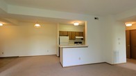 Located at 1040 Windward Dr in London, Ohio, this single-floor residence offers a practical and comfortable layout designed for easy everyday living. The open-concept living area flows seamlessly into the kitchen, which is defined by a peninsula-style pass-through counter that connects both spaces while maintaining an airy, open feel. The kitchen is equipped with warm wood-toned cabinetry, white appliances including a full-size refrigerator, a built-in range with overhead ventilation, and a sink positioned along the counter — all set beneath flush-mount ceiling lighting that keeps the space bright and functional. The living area features neutral wall-to-wall carpeting that adds warmth underfoot, and is complemented by a hanging pendant light that adds a subtle decorative touch. Clean white walls throughout contribute to a light and welcoming ambiance. The space is finished with standard trim details and ceiling vents for climate control, reflecting a straightforward, well-maintained interior. Overall, this single-story residence in London, OH presents a clean, move-in ready environment well-suited for those seeking a functional and comfortable home in a quiet neighborhood setting.