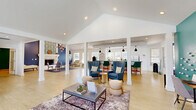 Located at Lakeshore Trail East in Indianapolis, Indiana, this generously sized single-floor living space offers a thoughtfully designed community gathering area that blends style with functionality. The space is defined by dramatic vaulted ceilings that rise to impressive heights, creating an open and airy atmosphere throughout. A striking navy blue accent wall frames an elegant white-mantled fireplace, topped with a bold patterned tile surround and complemented by an eye-catching geometric chandelier overhead. Light hardwood-style flooring runs throughout, unifying the various activity zones within the open floor plan. Tall windows and an arched transom window flood the interior with natural light, offering views of the surrounding outdoor areas. A lounge zone features high-back upholstered chairs in rich teal and blue tones arranged around accent tables, while a bar-height counter with vibrant green stools provides a casual seating area. A teal accent wall anchors another section of the space, showcasing a wall-mounted television above a wooden credenza. Crisp white columns add architectural structure without interrupting the open flow. Crown molding and clean white trim throughout lend a polished, classic finish to this vibrant and welcoming community space.