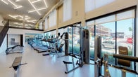 This fitness center is designed with both functionality and aesthetics in mind, offering a spacious, open layout beneath impressively high ceilings that lend the space an airy, energizing atmosphere. Geometric LED light fixtures suspended overhead provide bright, even illumination throughout, complementing the abundant natural light that streams in through large, floor-to-ceiling windows lining one wall. These expansive windows offer views to the outside, keeping the interior feeling connected to the surrounding environment.

The flooring throughout is a durable, light-toned surface well-suited for high-traffic fitness use. A full-length mirror runs along one wall, visually expanding the space while allowing users to monitor their form during workouts. The cardio zone features a lengthy row of treadmills, ellipticals, and stair climbers positioned along the windowed wall. Toward the interior, a dedicated strength training area houses cable machines, a power rack, adjustable benches, and a well-stocked dumbbell and kettlebell rack. A wall-mounted flat-screen television is positioned near the free weights for entertainment. A staircase with a sleek dark railing leads to an upper mezzanine level, adding additional dimension to the overall facility.