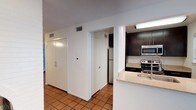 This single-floor space presents a practical and straightforward layout centered around a kitchen visible from the main hallway area. The kitchen is equipped with stainless steel appliances, including an electric range, overhead microwave, and a full-size refrigerator tucked into an alcove. Dark espresso-toned upper cabinetry contrasts cleanly against white walls, while a light-toned granite or stone countertop extends to form a peninsula with a built-in sink, offering a casual workspace or serving area. Recessed fluorescent lighting illuminates the kitchen efficiently. Terracotta ceramic tile flooring runs throughout the visible areas, adding warmth and a classic character to the space. The hallway adjacent to the kitchen features multiple white panel doors, likely leading to closets or utility areas, along with visible HVAC vents indicating a centralized climate control system. A wall-mounted electrical panel is also present in the corridor. The overall aesthetic is clean and utilitarian, with a neutral white palette complementing the darker kitchen elements and earthy floor tones.