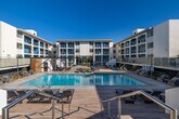 Located at 13900 Panay Way in Marina del Rey, California, this outdoor amenity space delivers a resort-caliber experience in one of Los Angeles County's most sought-after coastal communities. The centerpiece of the space is a generously sized swimming pool with crystal-clear turquoise water, framed by a wide, light-toned tile and wood-plank pool deck that invites relaxation throughout the day. Surrounding the pool, an array of cushioned chaise lounges and low-profile lounge chairs are thoughtfully arranged to maximize sun exposure and comfort. A pergola-style shade structure with warm wood detailing anchors one end of the deck, providing a shaded retreat with additional seating for groups or individuals seeking respite from the Southern California sun. Glass railings and wood slat privacy screens define the perimeter of the space, lending a modern, refined aesthetic while maintaining an open, airy feel. The deck is flanked on multiple sides by the multi-story residential building, which features a contemporary architectural design with arched balconies, white stucco facades, and large windows. Lush landscaping with olive trees and coastal plantings softens the hardscape and adds a natural, Mediterranean-inspired character. Under the clear blue skies of Marina del Rey, this outdoor space offers an exceptional blend of style, comfort, and California coastal living.
