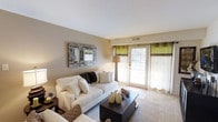 This single-floor space centers around a thoughtfully designed living room that balances comfort and style. The room features warm, neutral-toned walls complemented by plush, white upholstered seating arranged around a dark-finished rectangular coffee table, creating an inviting focal point. A decorative mosaic-framed mirror adds visual interest to the main wall, contributing an artistic touch to the otherwise understated palette.

Natural light is a defining feature of this living room, streaming in through a full-length glass door dressed with two-toned curtains in green and white, which introduce a subtle pop of color against the neutral backdrop. The door appears to lead to an outdoor area, enhancing the sense of openness and connectivity to the exterior. Soft carpeting underfoot adds warmth and comfort to the space.

Recessed ventilation is visible in the ceiling, and a floor lamp positioned near the seating area provides supplemental ambient lighting. The overall ambiance is calm and welcoming, with a cohesive design aesthetic that emphasizes clean lines, layered textures, and a harmonious neutral color scheme throughout.