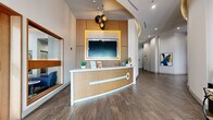 This single-floor lobby and common area serves as the welcoming centerpiece of the Radius residential community. The space is thoughtfully designed with a contemporary aesthetic, anchored by a sleek, curved reception desk finished in warm wood tones and crisp white accents. A yellow-framed flat-screen display mounted on a wood-paneled feature wall adds a bold pop of color behind the desk, complemented by a striking cluster of metallic globe pendant lights overhead.

The flooring throughout features large-format wood-look tile laid in a diagonal pattern, lending a sense of expansiveness and visual flow. A wide corridor connects the entrance — framed by tall glass doors that flood the space with natural light — to the elevator lobby, where a branded runner, cove-lit curved ceiling, and bold abstract artwork create a polished, gallery-like feel.

The lounge area transitions naturally from the reception zone, offering plush blue sofas and armchairs alongside intimate wood-paneled booth seating. Wall-mounted televisions and curated geometric artwork in blue tones enhance the relaxed yet refined atmosphere. Floor-to-ceiling windows with sheer printed curtains bathe the seating area in natural light. The overall ambiance is modern, welcoming, and thoughtfully curated throughout.