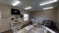 This single-floor multipurpose room is designed to accommodate a variety of social and recreational activities. The space features neutral gray walls and dark flooring, creating a clean, understated backdrop that complements its eclectic furnishings. Overhead, fluorescent panel lighting provides bright, even illumination throughout the room.

A wall-mounted flat-screen television serves as a focal point on one wall, flanked by decorative film reel accents and circular wall art, lending the space a casual, cinema-inspired character. A white console table beneath the TV offers surface space for décor and small items.

The seating arrangement includes a mix of white wooden chairs and a slatted coffee table, offering a relaxed and flexible setup. Folding chairs are stacked along one wall, suggesting the room can be reconfigured to suit larger gatherings or events. A tall bistro table with a bar stool in the corner adds an additional informal seating option.

A window fitted with horizontal blinds allows natural light to filter in. The overall ambiance is casual and adaptable, making this room well-suited for community screenings, small group meetings, or social gatherings.
