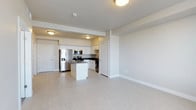 This single-floor space offers a clean, contemporary design centered around an open-concept layout that seamlessly connects the kitchen and living area. The kitchen is a standout feature, outfitted with stainless steel appliances including a French door refrigerator with an ice and water dispenser, a microwave, a range, and a dishwasher. White cabinetry with dark countertops provides a crisp contrast, while a central kitchen island adds both prep space and visual definition to the room. Light-toned flooring runs throughout the space, enhancing the bright and airy atmosphere created by flush-mount ceiling lighting. The open living area is generously proportioned, offering a blank canvas that accommodates a variety of furniture arrangements. Neutral tones on the walls and ceilings contribute to a calm, versatile ambiance. Entryway doors are positioned conveniently to provide privacy and separation from the main living space. The overall design prioritizes functionality and modern aesthetics, making it a well-suited space for everyday living.