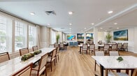 Located at 4824 Walmsley Blvd in Richmond, Virginia, this single-floor dining and kitchen space offers a welcoming and thoughtfully designed environment. The expansive dining area features warm light wood flooring, classic wainscoting along the walls, and large windows dressed with elegant drapes that flood the room with natural light. Multiple dining tables with marble-top surfaces and upholstered chairs are arranged throughout, accommodating both intimate gatherings and larger group settings. Bold, large-scale landscape artwork adorns the white walls, adding color and visual interest to the refined interior. Tall decorative trees and lush greenery punctuate the space, enhancing its vibrant atmosphere. The adjoining kitchen is efficiently laid out with dark wood cabinetry, white quartz countertops, and a classic subway tile backsplash. A striking teal accent wall above the cabinetry is complemented by framed artwork and trailing potted plants. Stainless steel appliances — including a full-size refrigerator, dishwasher, microwave, and a Keurig coffee station — provide comprehensive functionality. Recessed lighting throughout ensures consistent, well-distributed illumination. Together, the dining room and kitchen form a cohesive, inviting space that balances practicality with a warm, curated aesthetic.